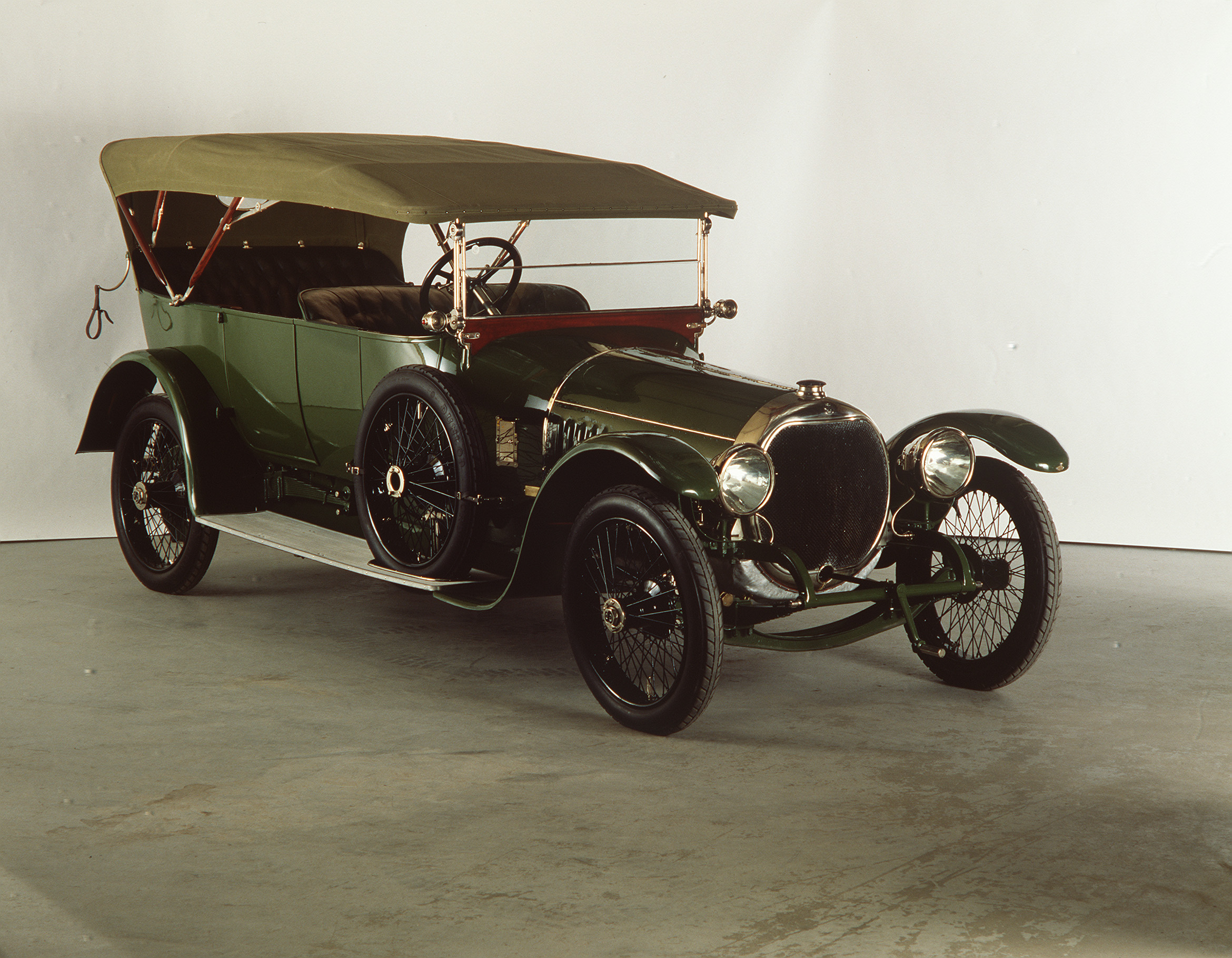 A black and green 1910s-era car with an open-top carriage and convertible roof-covering. The wheels of the car are very narrow yet have a wide diameter giving the car a high-set seating carriage.